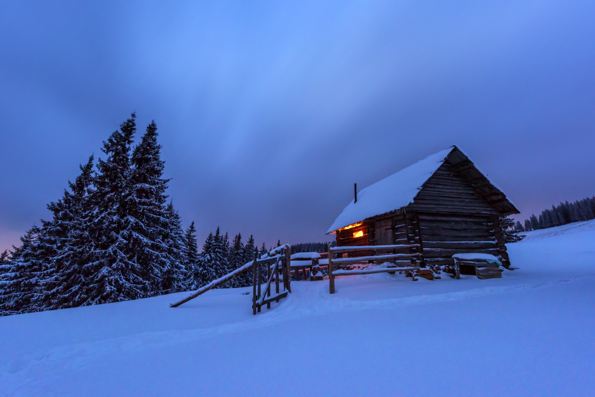 Snowy cabin in West Chester, PA, at dusk. Rustic wooden structure with light glowing from inside, surrounded by snow-covered landscape and pine trees.