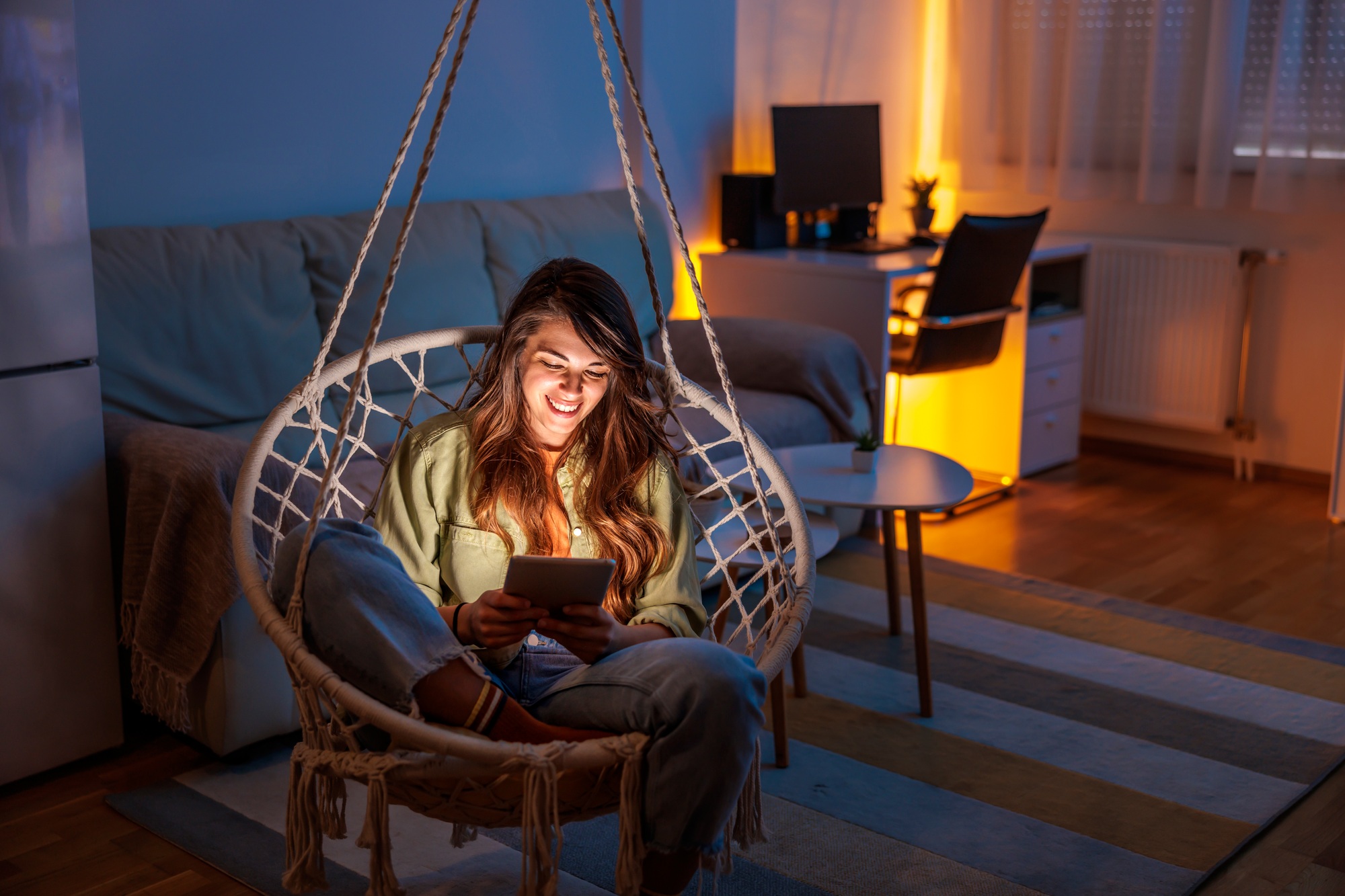 Woman relaxing in a hanging chair at home, illuminated by the light from her tablet. Comfortable living space. Interior design inspiration.