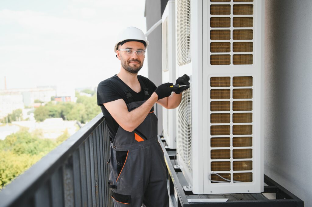 Air Conditioning Maintenance: Technician repairing an AC unit on a balcony. Man in West Chester, PA, wearing safety gear and using a screwdriver.